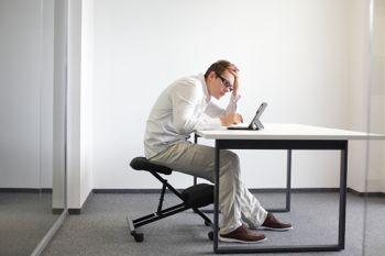 Young man is bent over her tablet in his office,seating on kneeling chair. Bat sitting posture at work.
